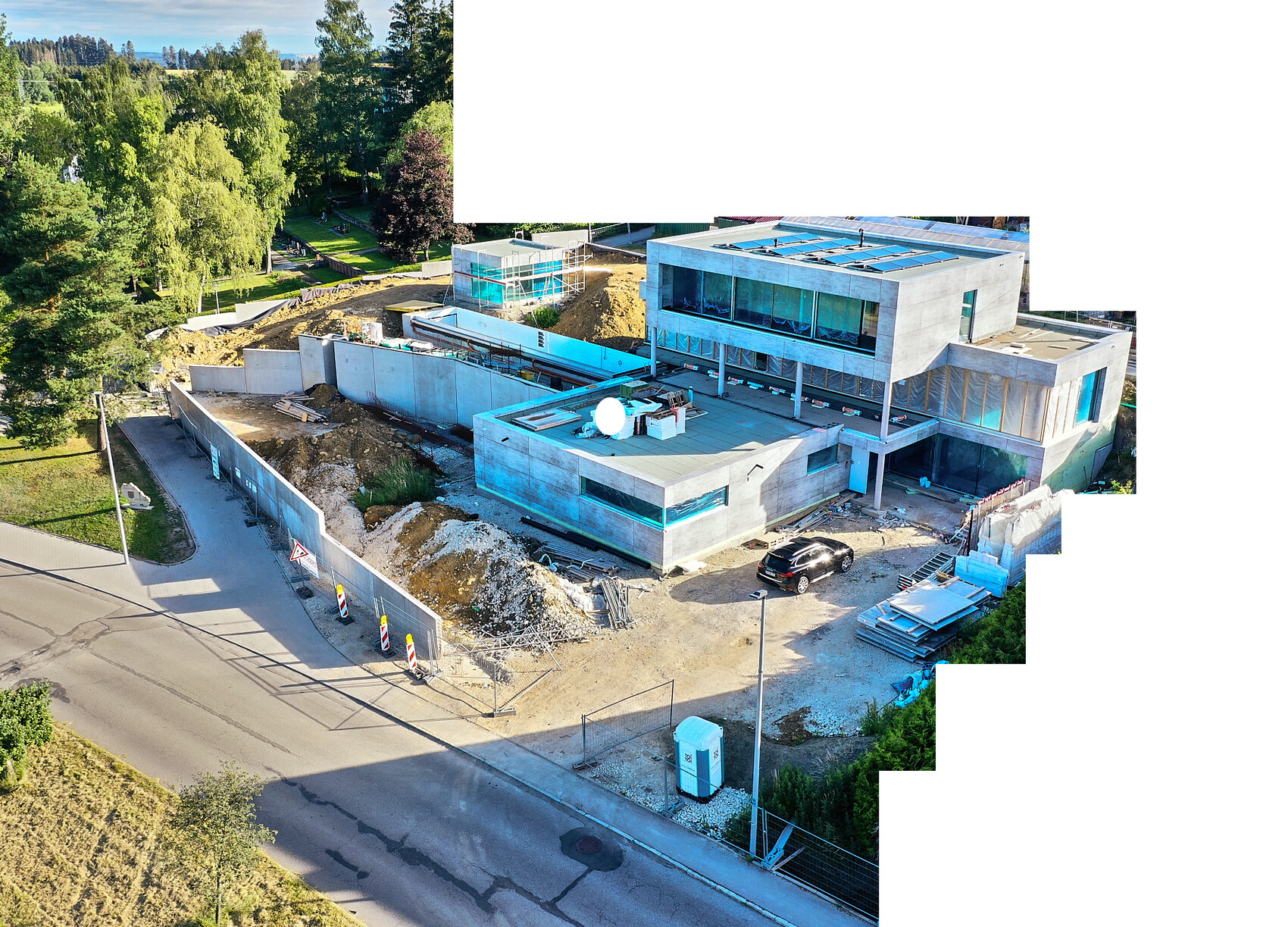 Luftaufnahme eines modernen Gebäudekomplexes im Bau: Betonwände, Glasfassaden, Bauzäune und Baugeräte, umliegende Grünflächen.