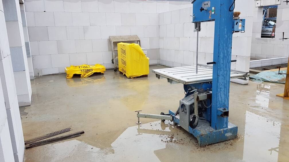 Nasse Innenbaustelle mit blauer Hebevorrichtung, gelben Säcken und Baumaterial.