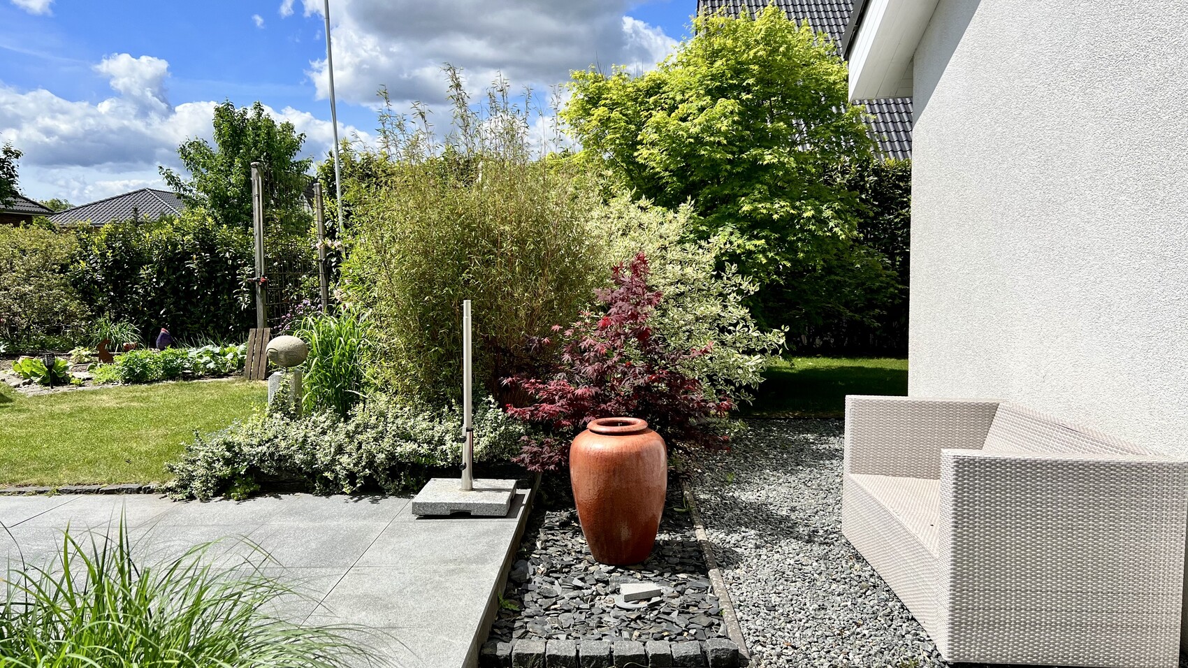 Garten mit Kiesweg, beige Sitzmöbel, orange Tonvase, grüne Sträucher, weiße Hauswand.