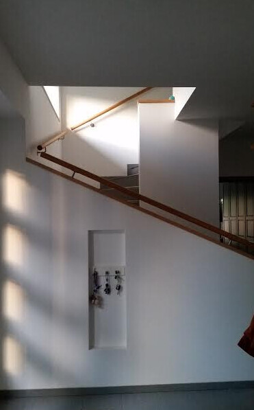 Heller Innenraum mit Treppe und diagonalem Holzhandlauf; weiße Wände, Nische mit Schlüsselhaken.