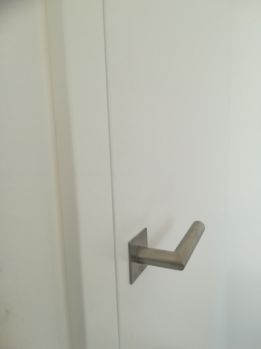Gebürsteter Edelstahl-Türgriff mit quadratischer Rückplatte an weißer Tür, seitliche Ansicht.