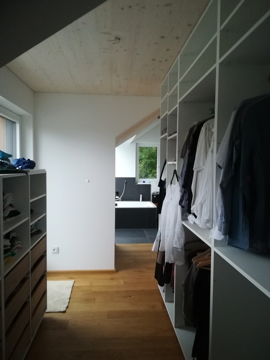 Garderobe mit offenen Regalen voller Kleidung, Holzboden und Blick auf Bad am Ende