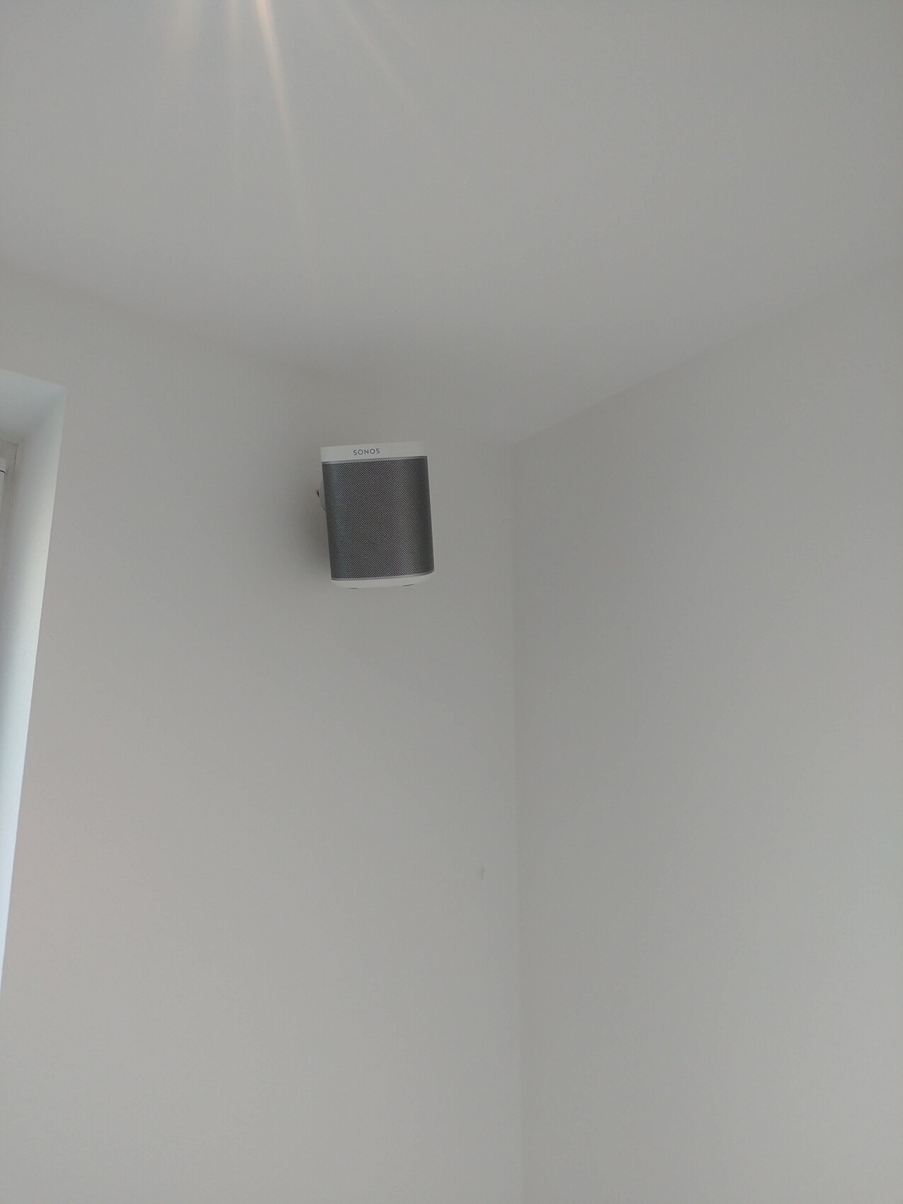 Weiße Zimmerwand-Ecke mit an der Wand montiertem Sonos-Lautsprecher in grauem Stoff.
