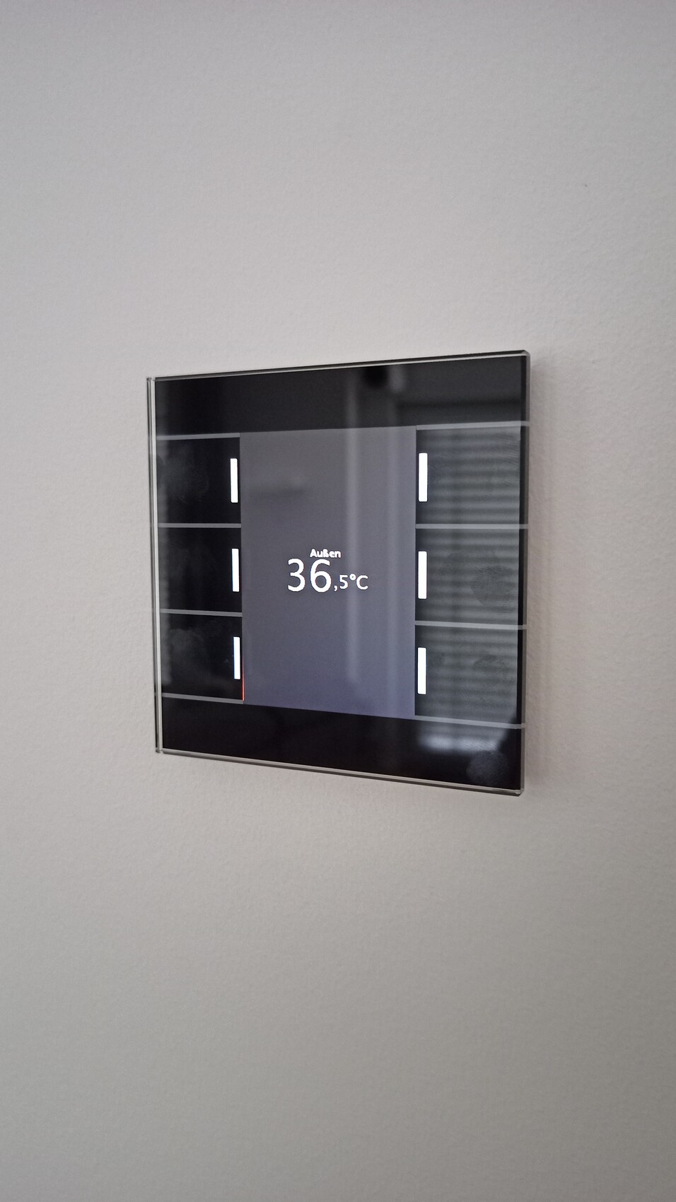 Wandmontiertes Smart-Thermostat mit Glasdisplay zeigt Außentemperatur 36,5°C.