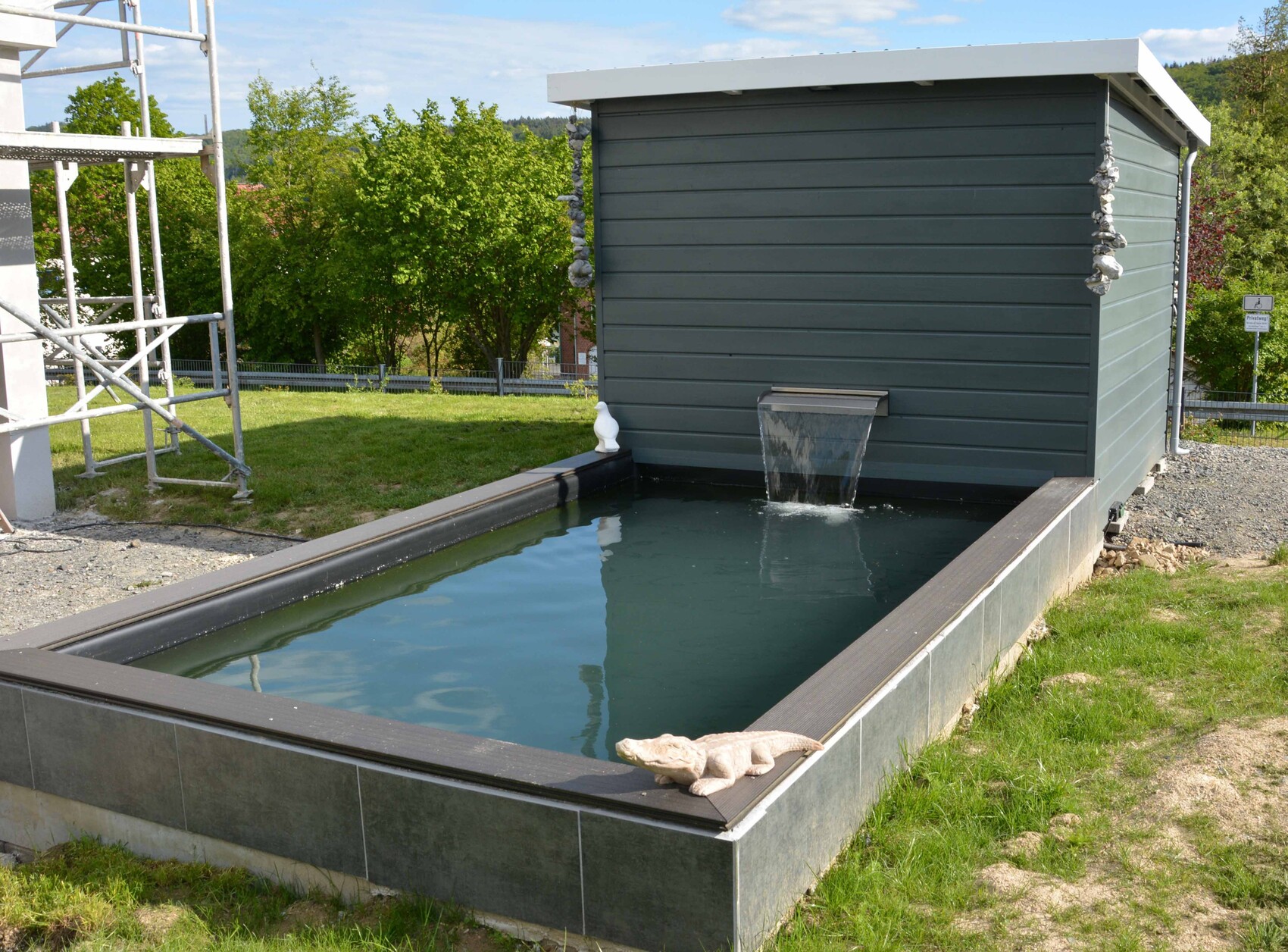 Rechteckiges Wasserbecken mit Wasserauslauf vor grauem Gartenhaus; Bäume dahinter.