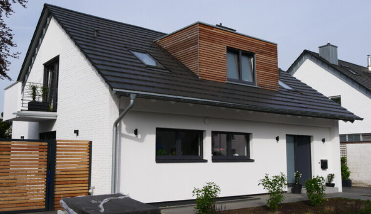 Modernes weißes Einfamilienhaus mit dunklem Satteldach, Dachgaube aus Holz und Garten.