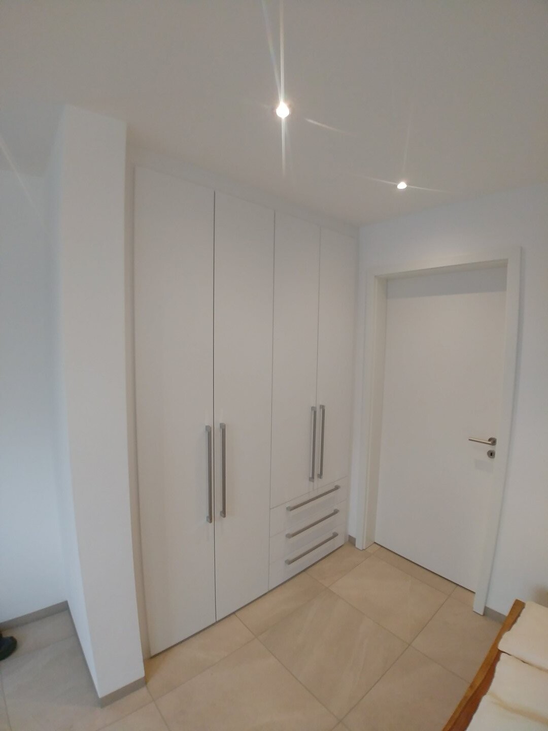 Weißer Kleiderschrank mit vier Türflügeln und drei Schubladen in hellem Raum; Tür rechts.
