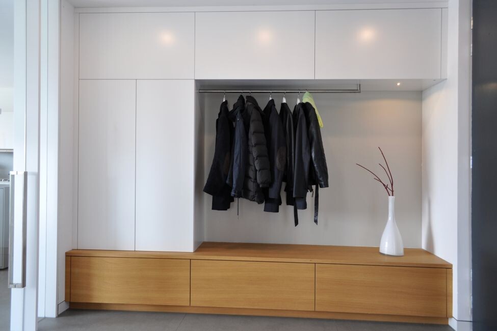 Moderne offene Garderobe: weiße Schränke, Hängestange, Jacken, Holzbank, Vase mit Zweigen.
