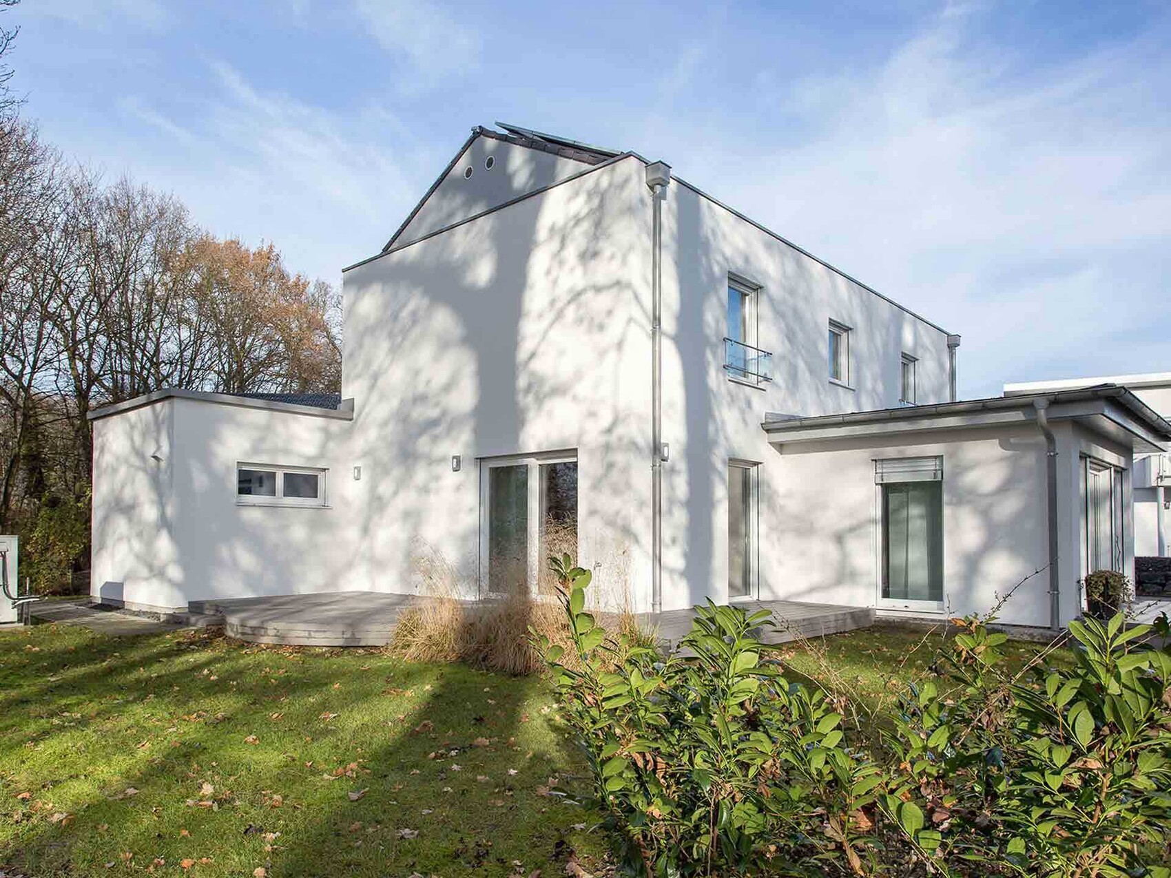 Modernes, weißes Einfamilienhaus mit Terrasse, großen Fenstern, Garten und Schatten von Bäumen.