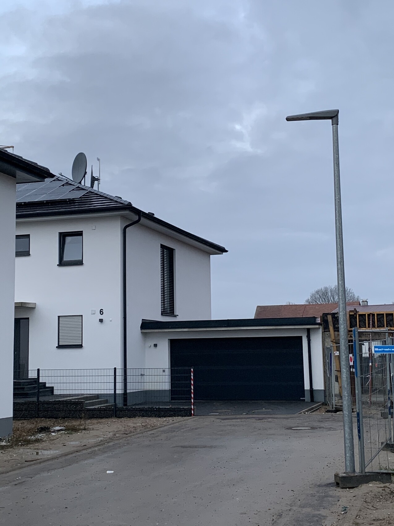 Modernes weißes Haus mit schwarzer Garage, Solarmodule und Satellitenschüssel; Bauzaun rechts