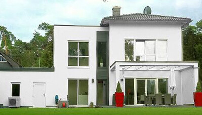 Moderne weisse Hausfassade mit grosser Terrasse und gruenem Garten im Vordergrund.