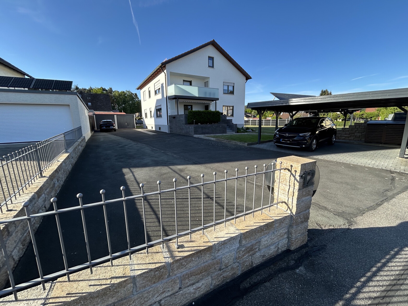 Modernes weißes Haus mit Carport rechts, schwarzem SUV, gepflasterter Einfahrt und Zaun.