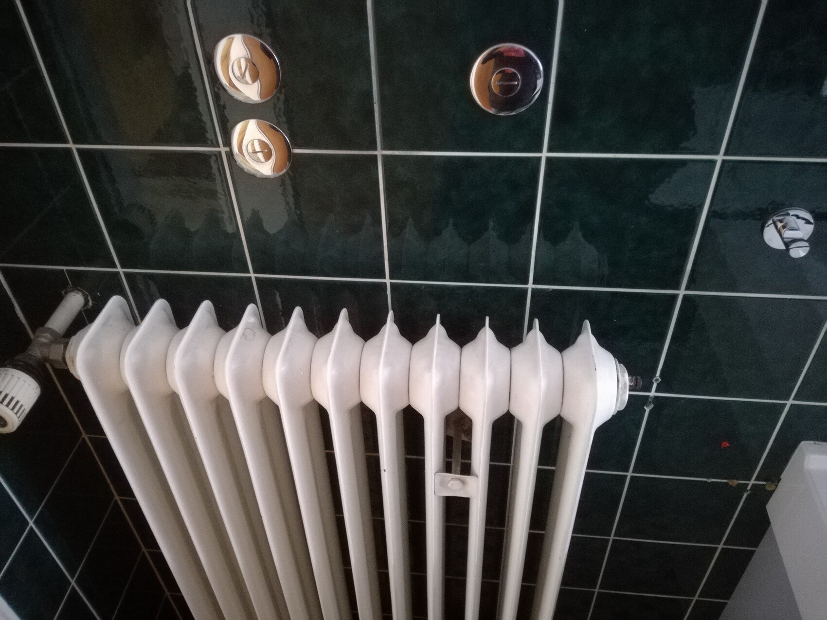 Weißer mehrteiliger Heizkörper vor dunkelgrüner Fliesenwand, mit sichtbaren Ventilen.