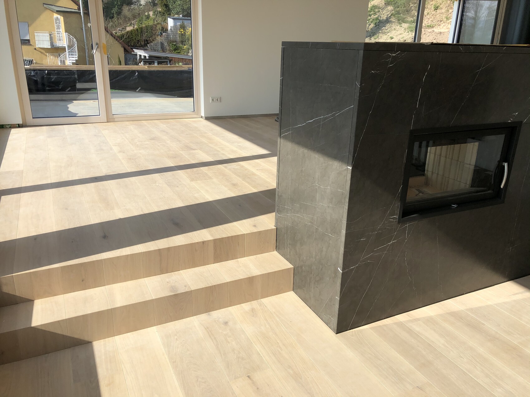 Heller Innenraum mit Holzboden, Treppenstufen, großen Glasfronten und dunklem Marmorkamin.