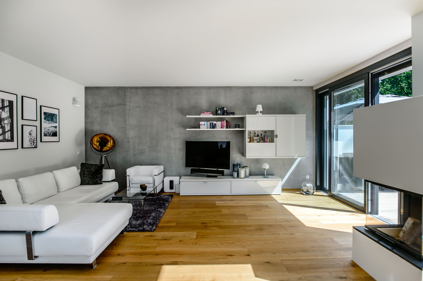 Helles Wohnzimmer mit weißer Couch, Betonwand, TV-Unit, Regale und Glasfront nach draußen.