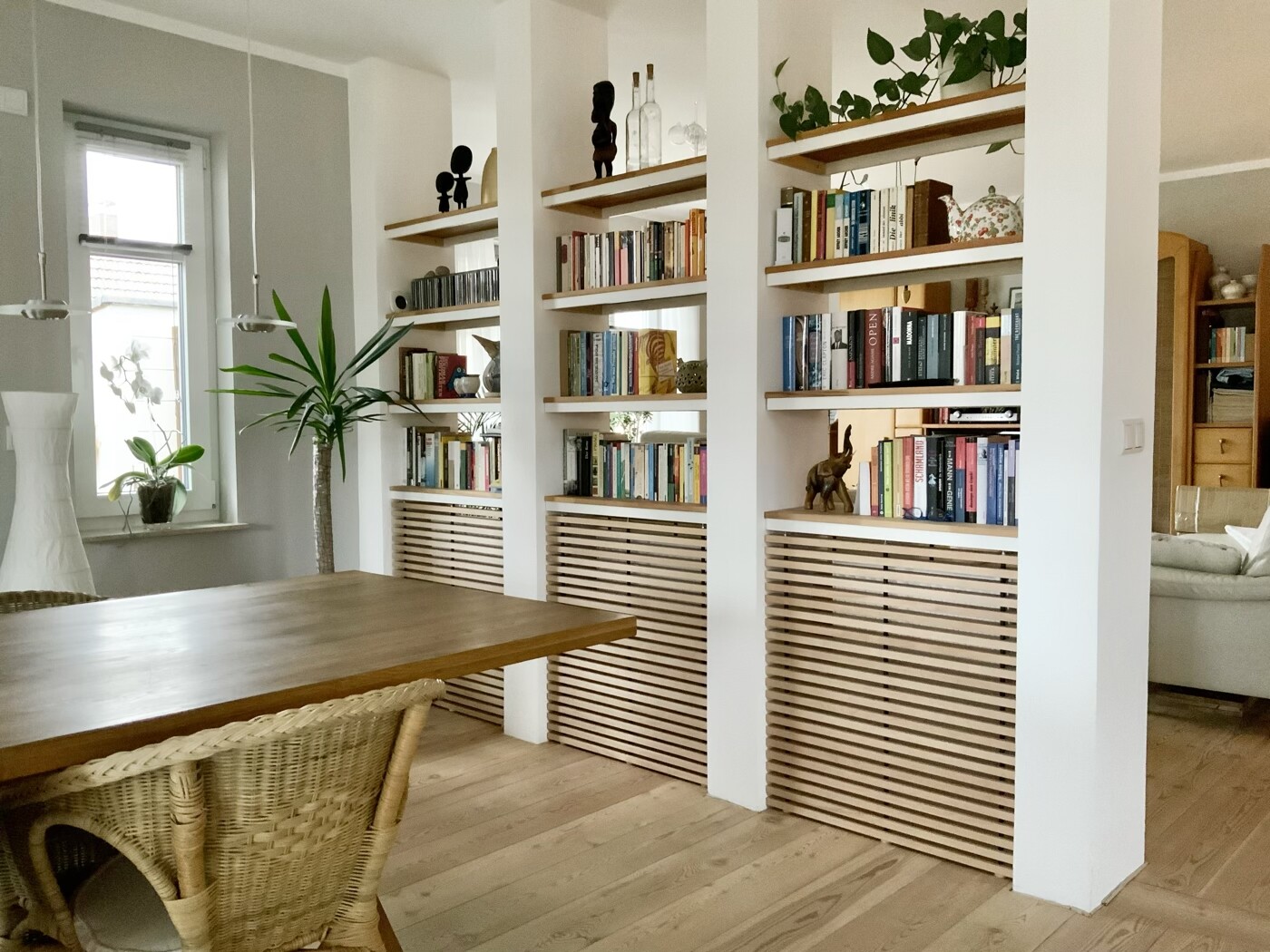 Heller Wohnraum mit durch Säulen geteilten Holzregalen voller Bücher, Pflanzen und Holztisch.