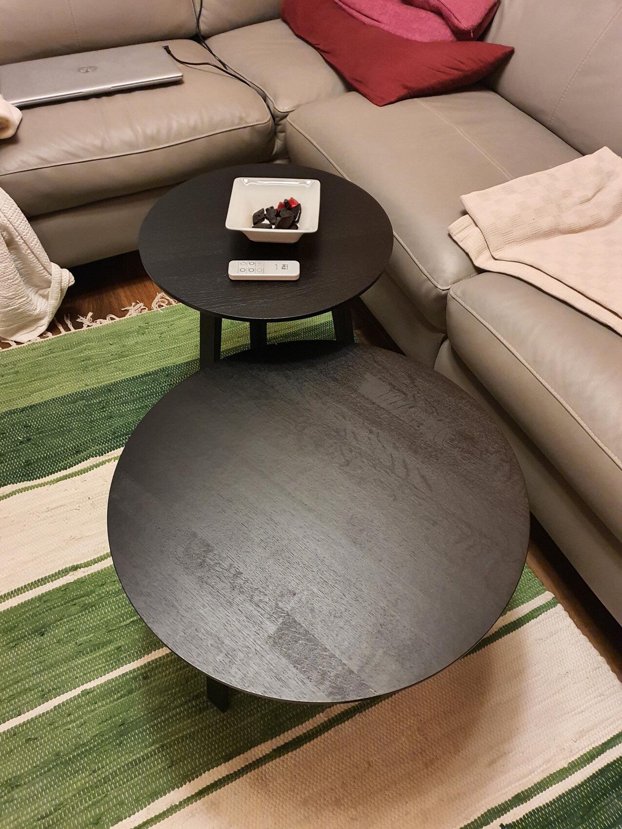 Zwei runde schwarze Tische vor grauer Couch; Schale mit dunkler Schokolade, Fernbedienung.