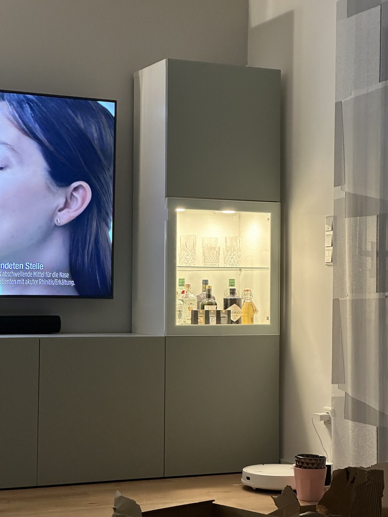 Wohnzimmer: links Flachbildfernseher, beleuchtete Glasvitrine mit Getränken, graue Schränke.