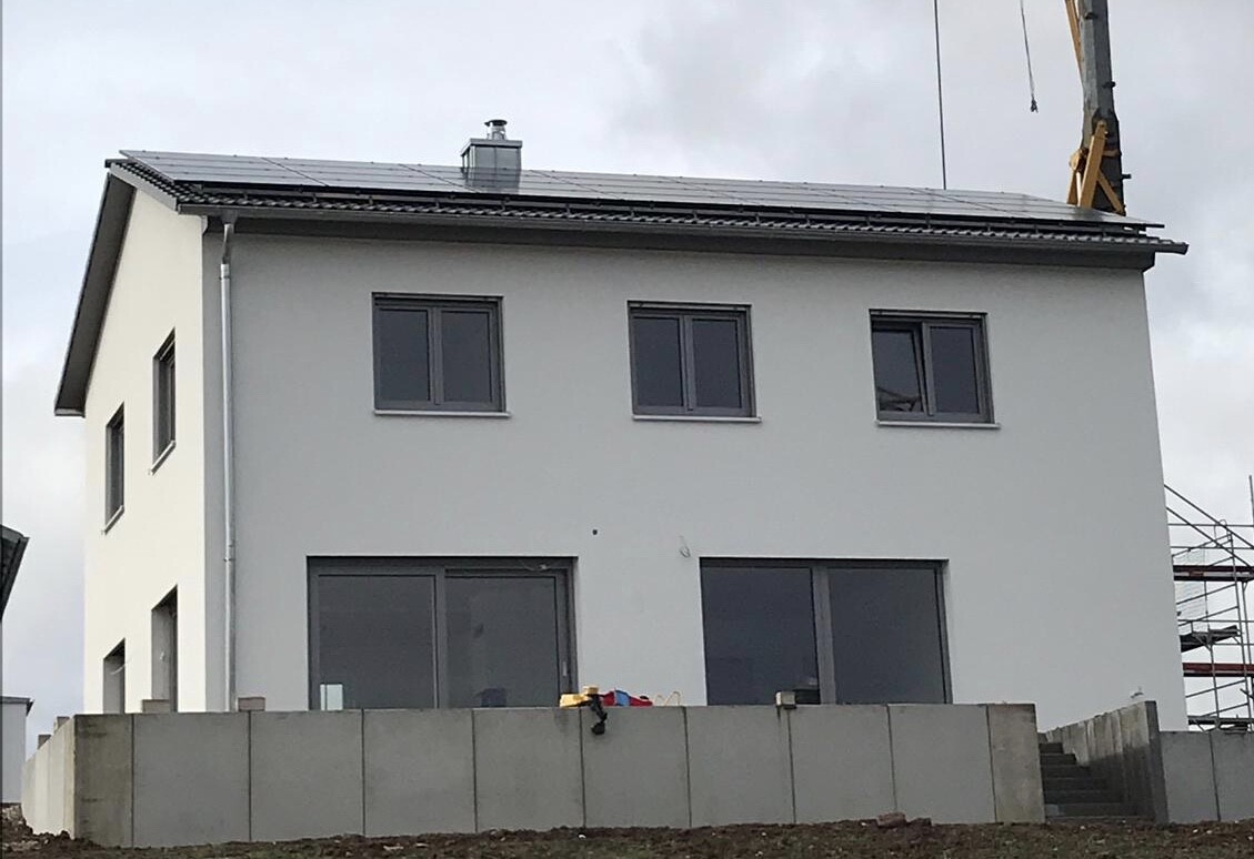 Weißes zweistöckiges Haus mit Solarmodulen auf dem Dach und Betonmauer davor.