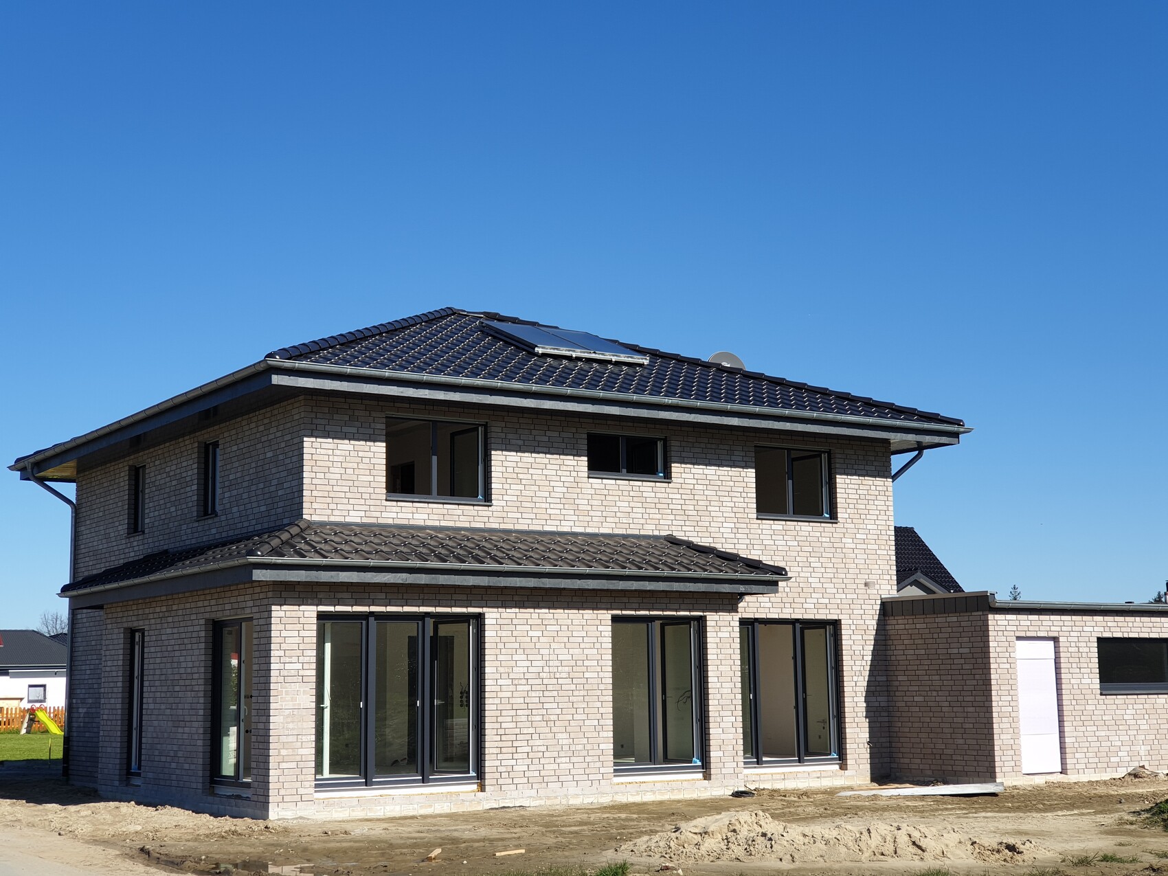 Modernes zweistöckiges Ziegelhaus mit dunklem Dach und Solarpanelen; Baustellenbereich davor.