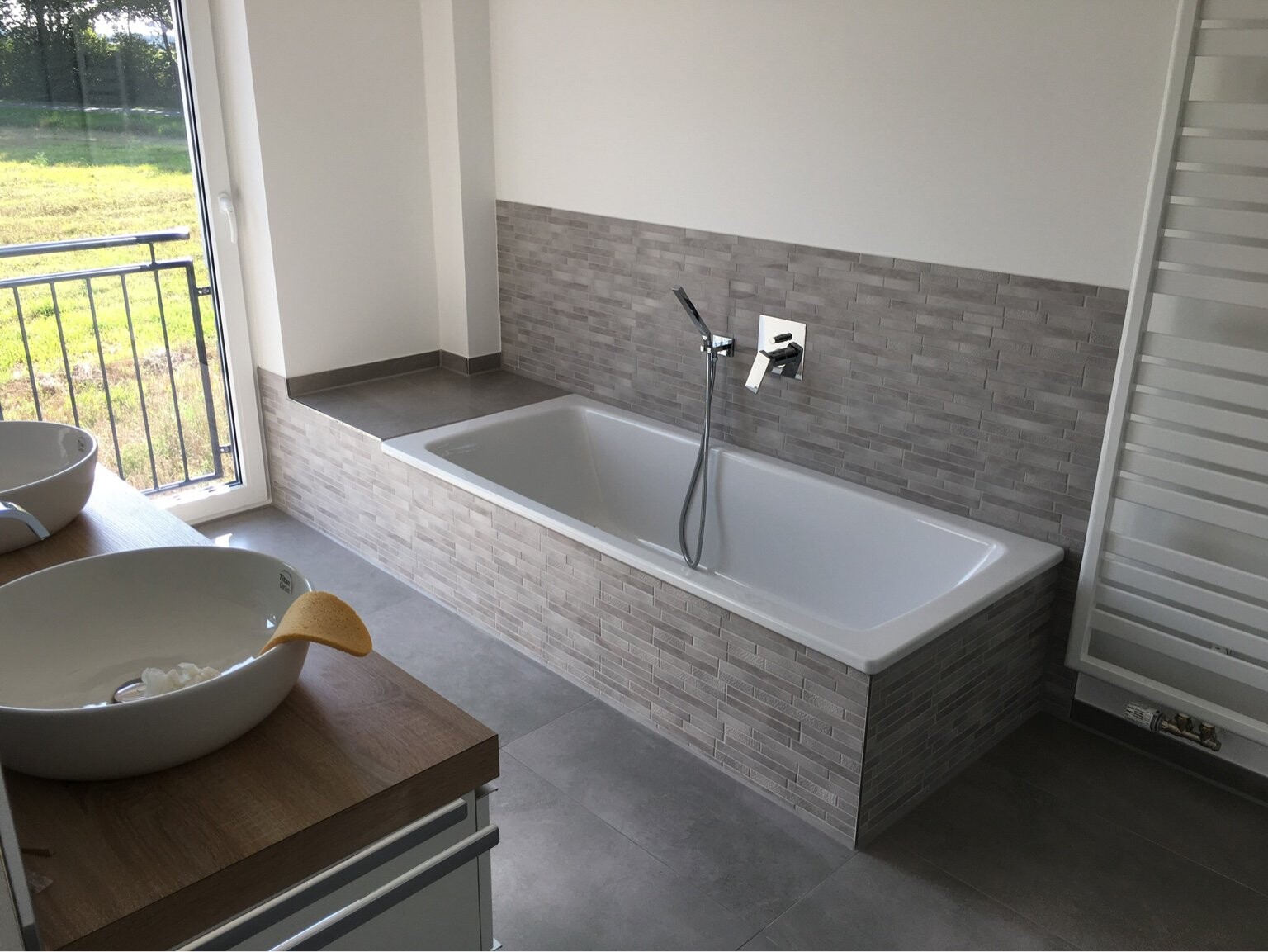 Modernes Badezimmer mit weißer Badewanne an grauer Fliesenwand, Fenster nach draußen, Holz-Waschtisch.