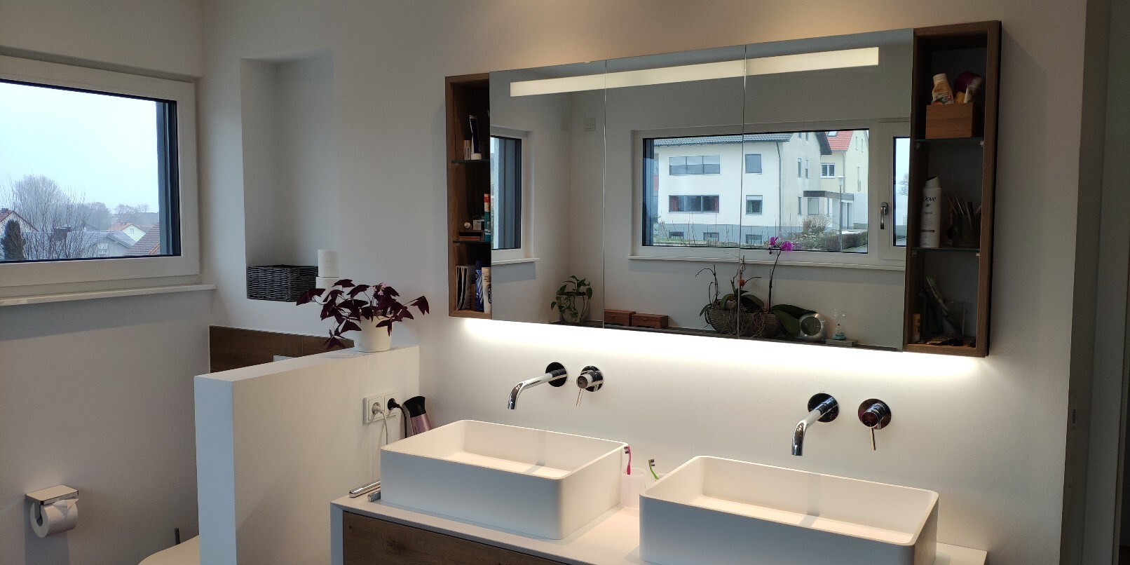 Modernes Badezimmer mit Doppelwaschbecken, beleuchtetem Spiegel, Ablagen und Fensterblick.