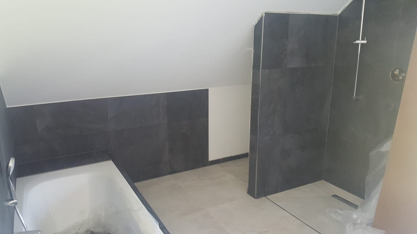 Dunkles Badezimmer mit schwarzer Fliesenwand, Badewanne links, Dusche rechts.
