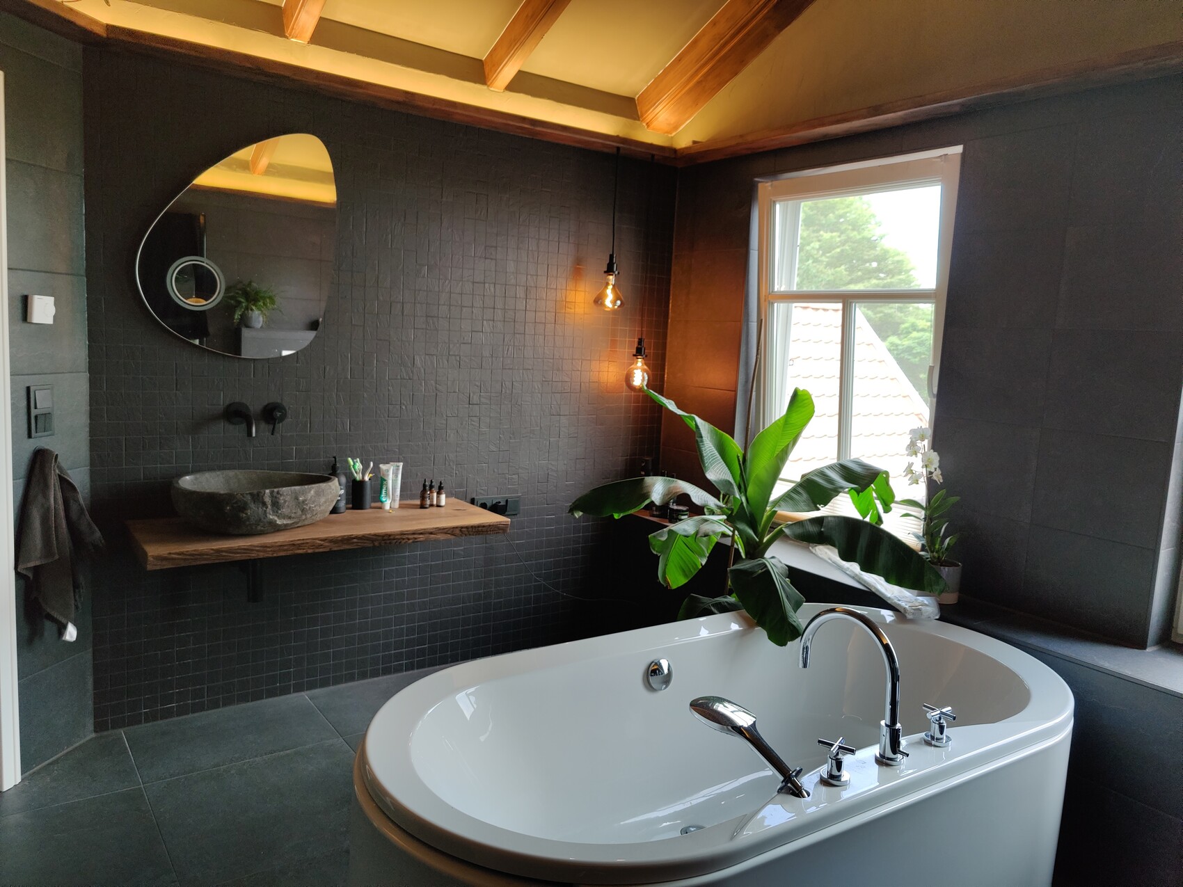 Freistehende Badewanne vor dunklen Fliesen; Holzregal mit Steinbecken, runder Spiegel, Pflanze.