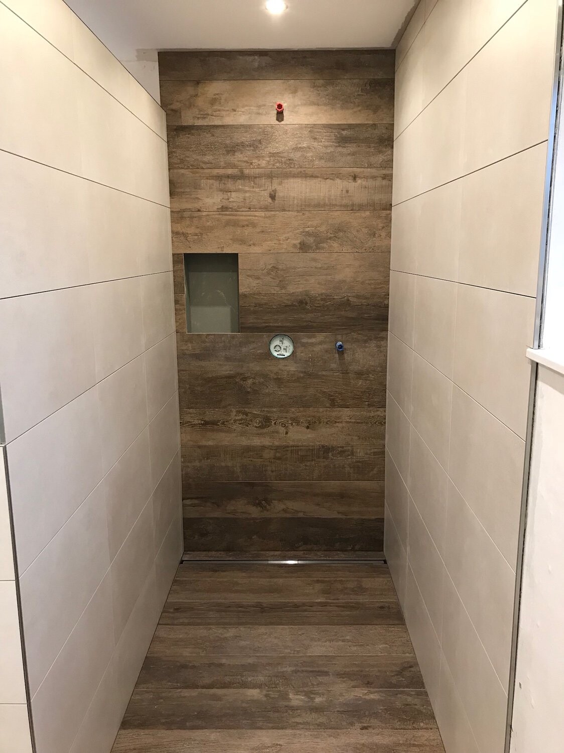 Dusche mit Holzoptik-Rückwand, seitliche weiße Fliesen, rotes Ventil und Zähler.