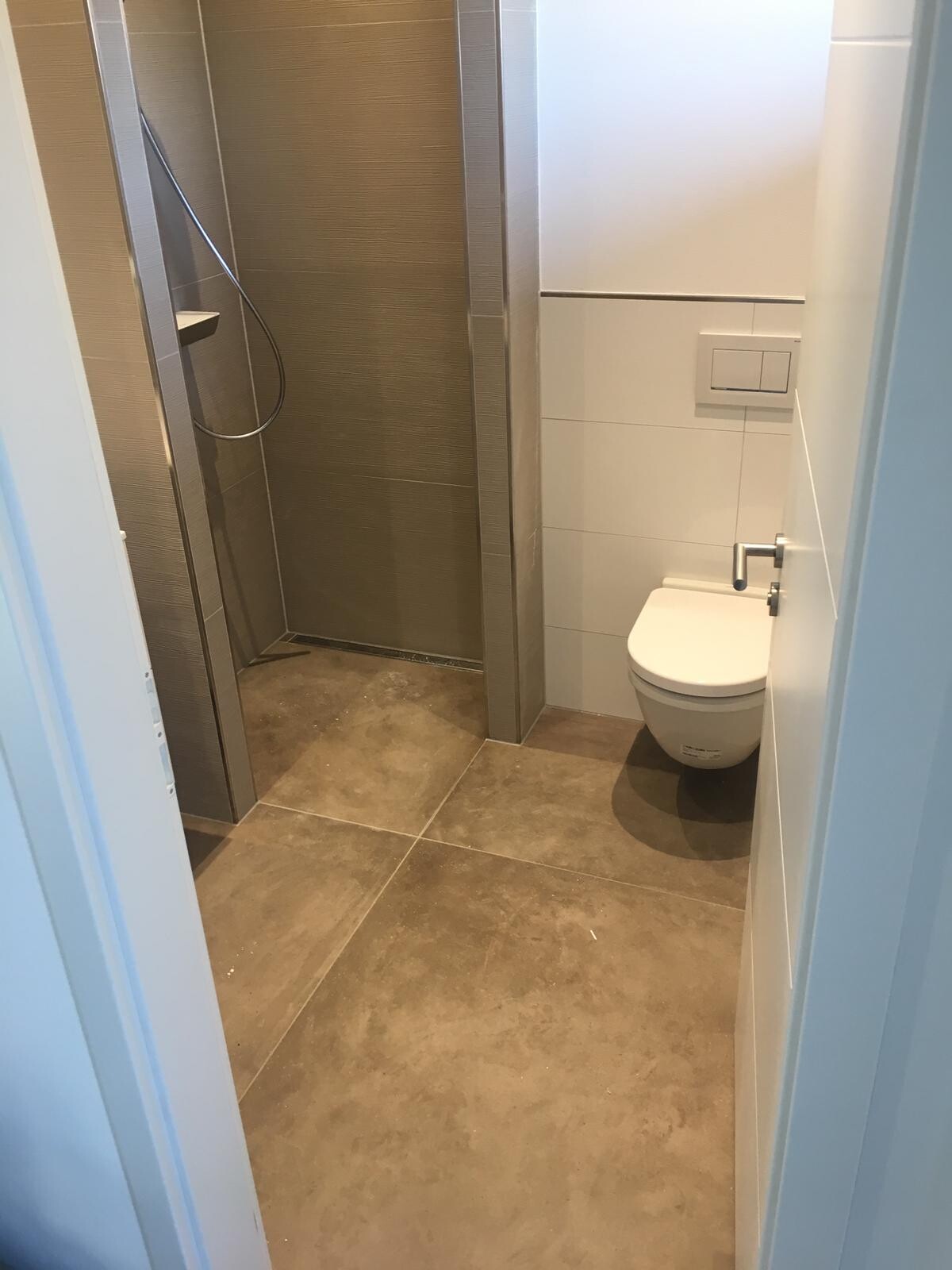 Kleines Badezimmer: Dusche links, wandhängendes WC rechts, graue Boden- und braune Wandfliesen.
