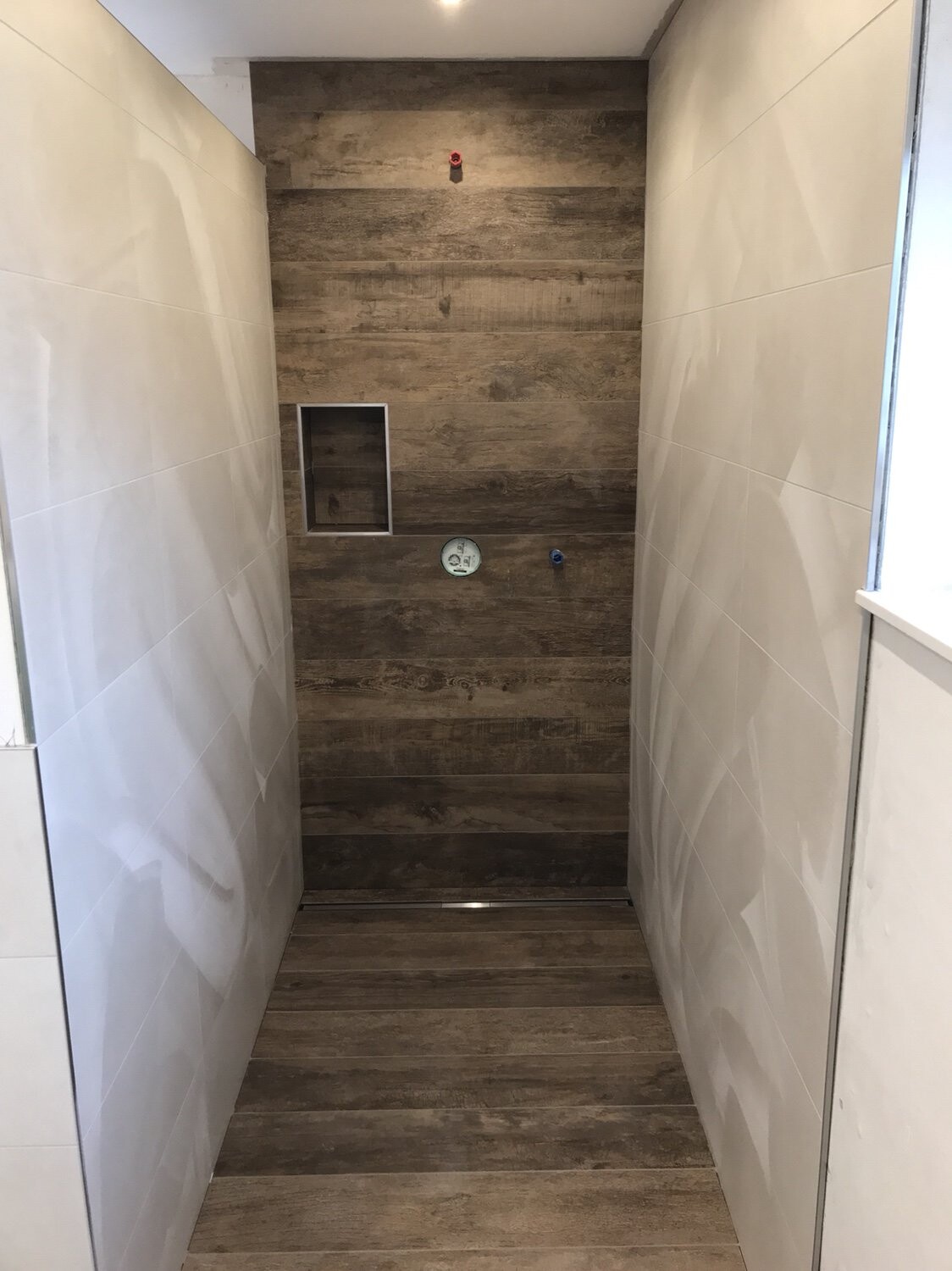Enge Dusche mit dunkler Holzwand, weiße Seitenfliesen, linke Nische und rundes Ziffernblatt in Mitte.