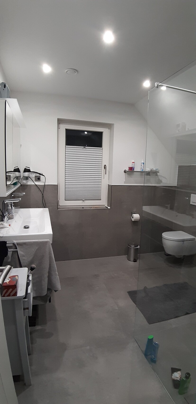 Modernes Badezimmer mit grauen Fliesen, Glasdusche, WC, Doppelwaschbecken und Fenster mit Rollladen.