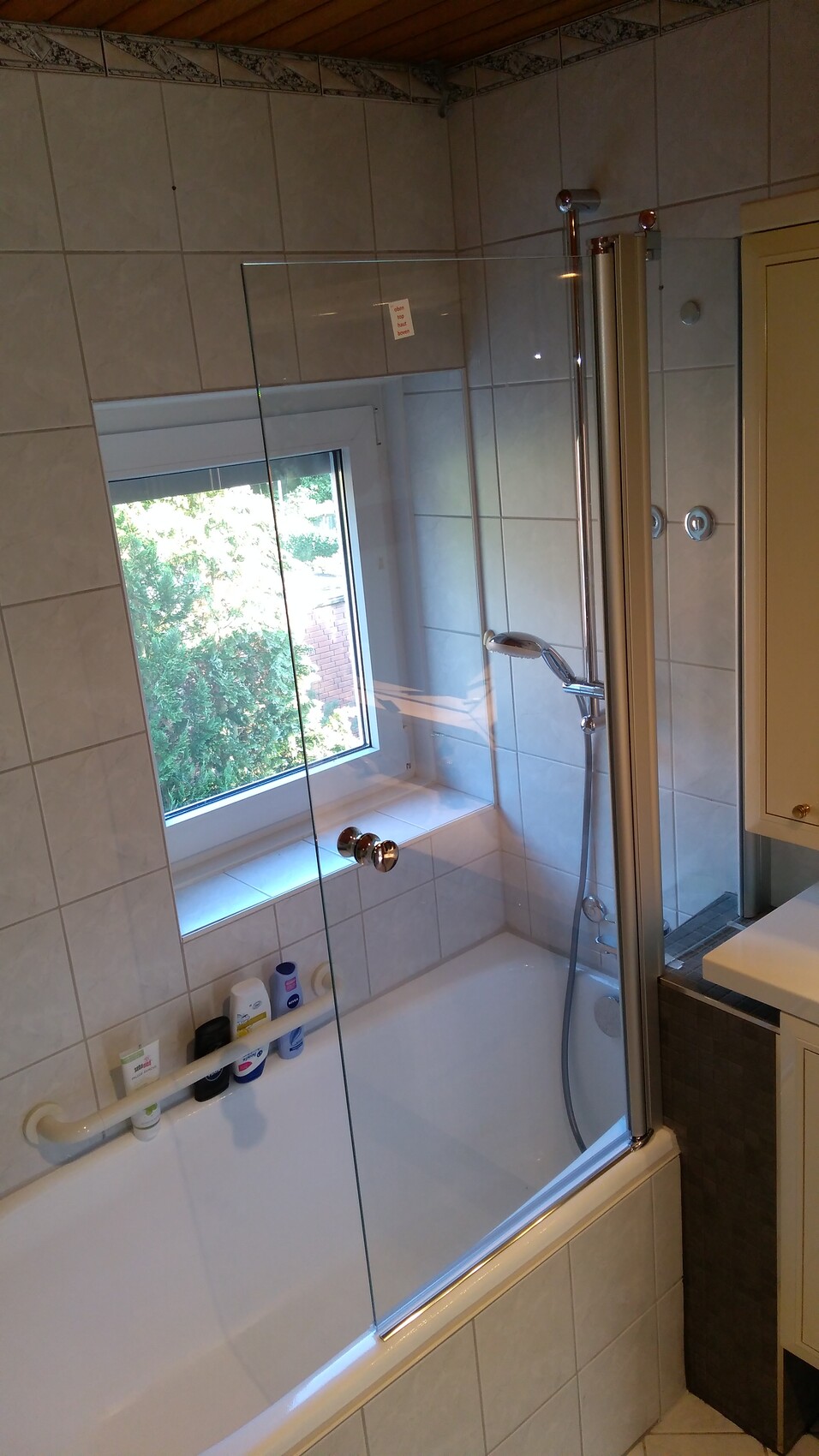 Badezimmer mit Badewanne, Glastür-Duschabtrennung, Fenster und Hygieneprodukten am Rand.