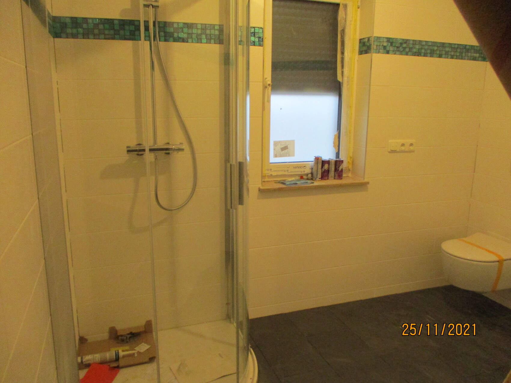 Badezimmer mit Glasdusche links, Fenster mittig, Toilette rechts; Baustellenmaterial auf dem Boden.