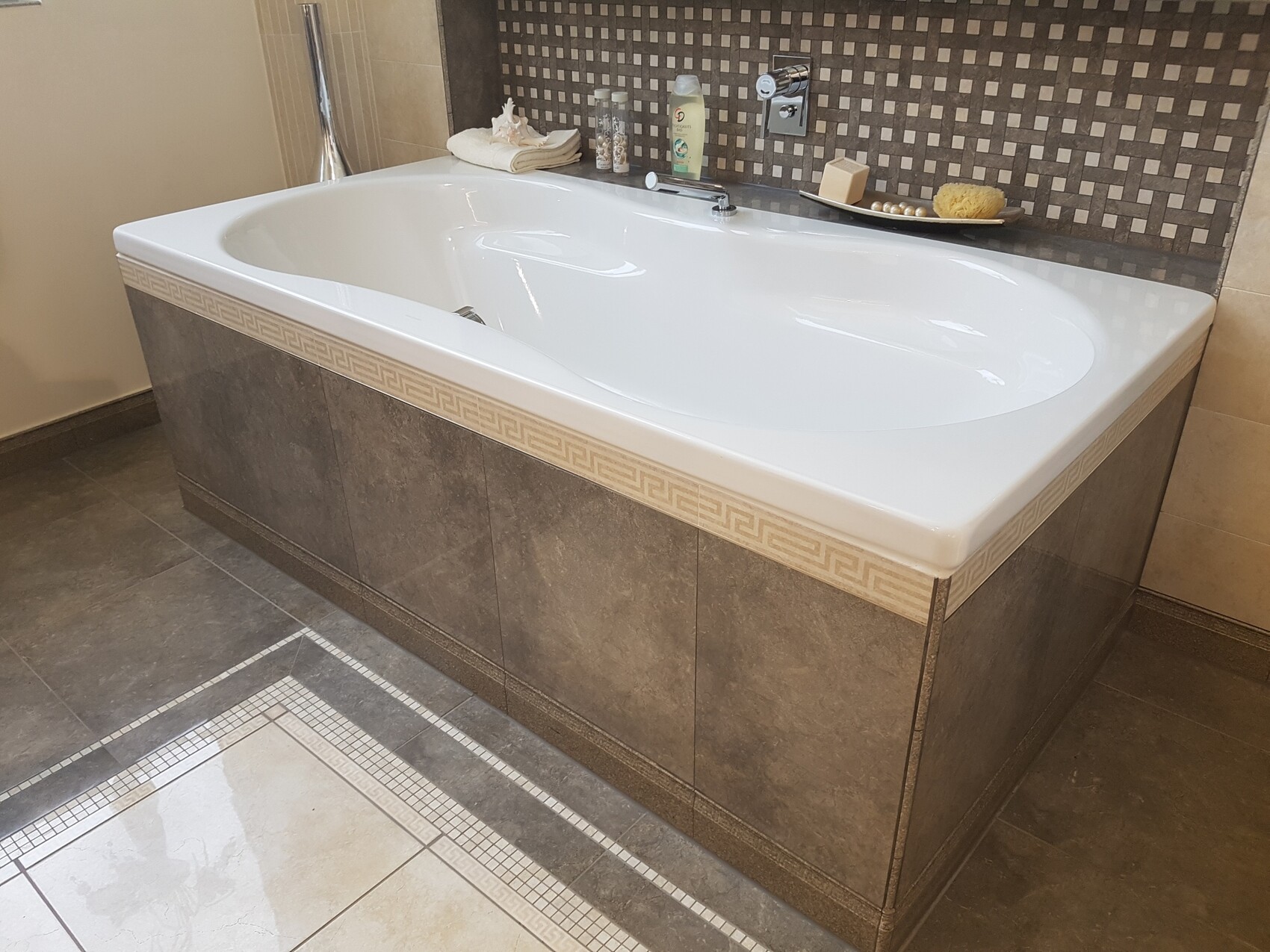 Große weiße Badewanne vor gemusterten grauen Fliesen; Badutensilien liegen am Rand.