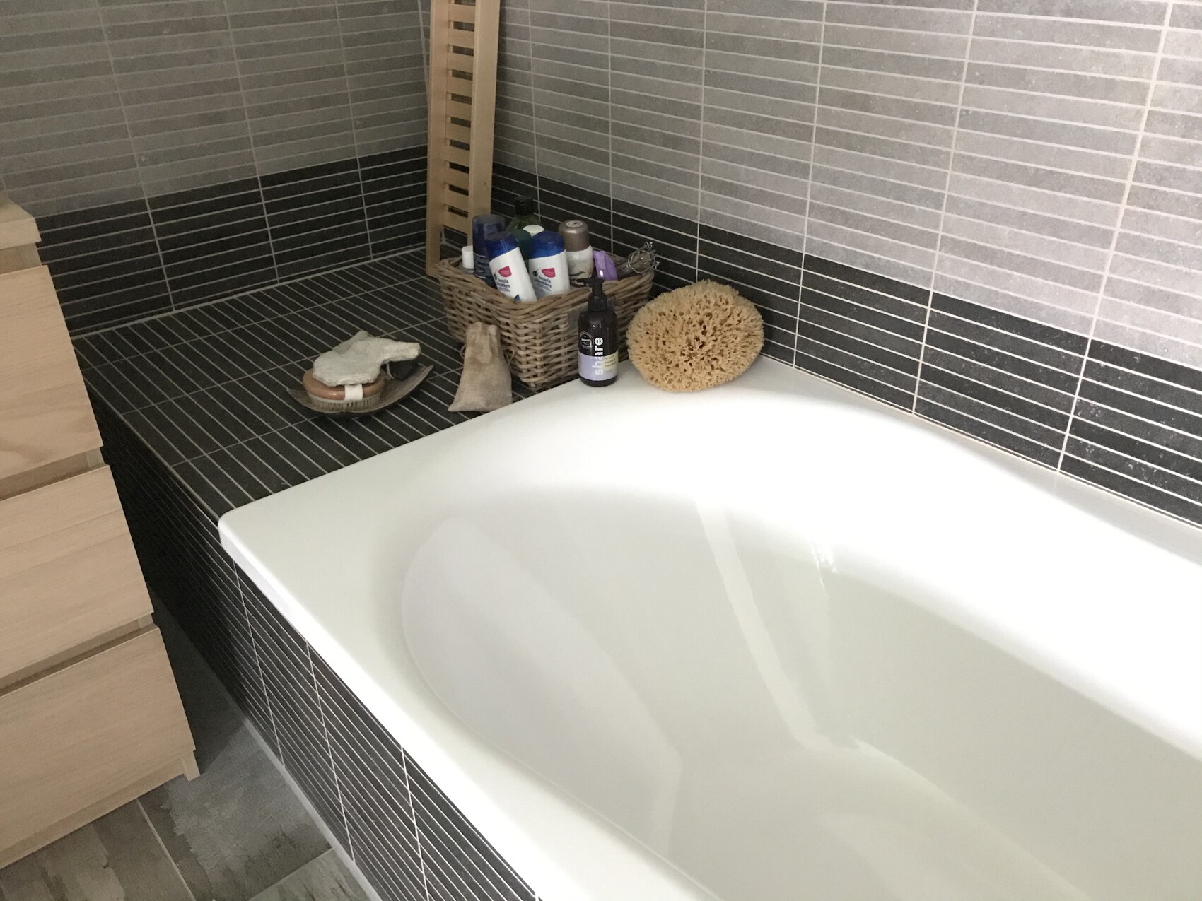 Badezimmer: weiße Badewanne, dunkle Fliesen, Korb mit Pflegeprodukten, runder Schwamm.