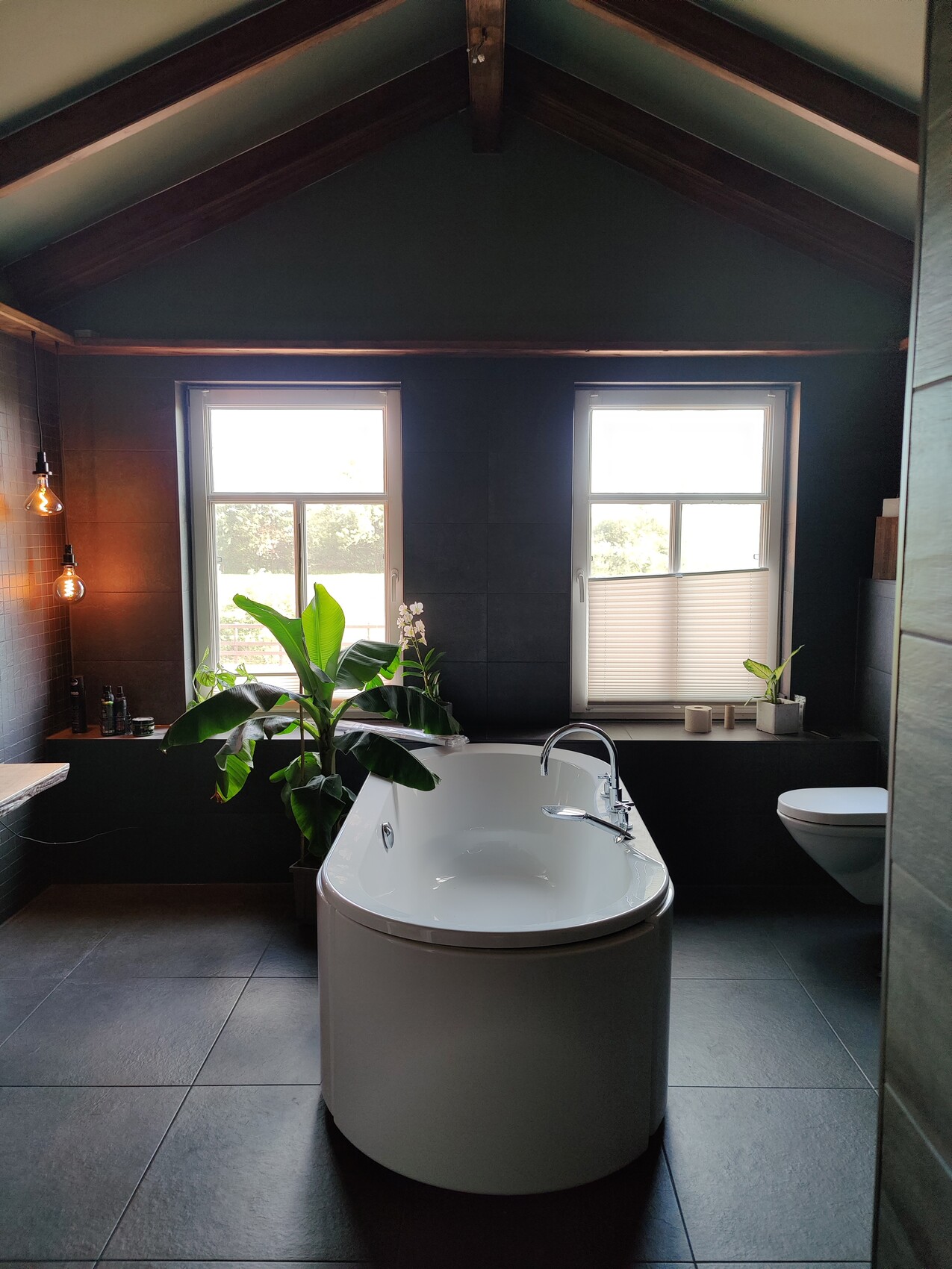 Helles Badezimmer mit freistehender Badewanne, dunklen Fliesen, mit zwei Fenstern und Pflanzen.
