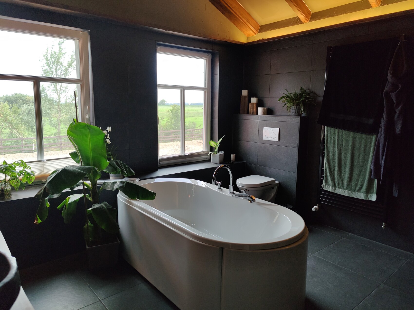 Freistehende weiße Badewanne vor dunklen Fliesen, große Fenster und Grünpflanzen.