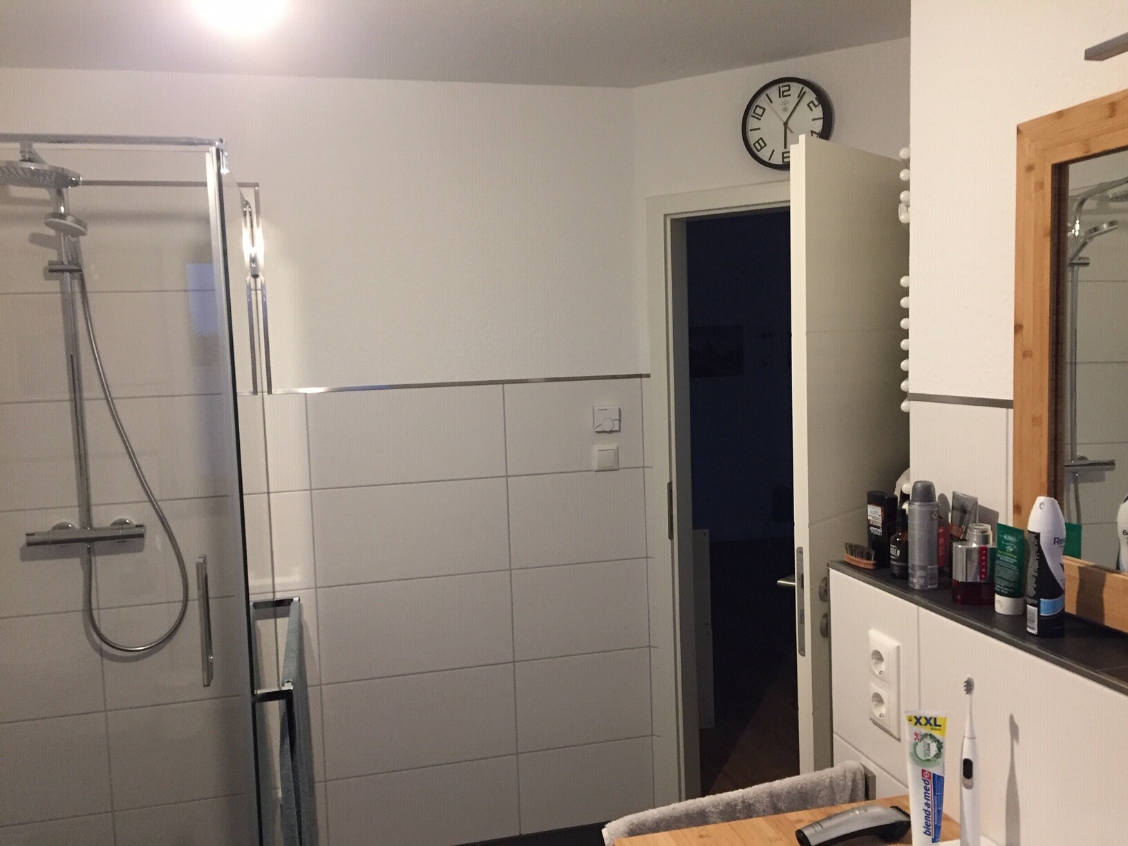 Badezimmer mit Glasdusche links, offene Tür zum dunklen Flur, Uhr über Tür; rechts Spiegel und Pflegeprodukte.