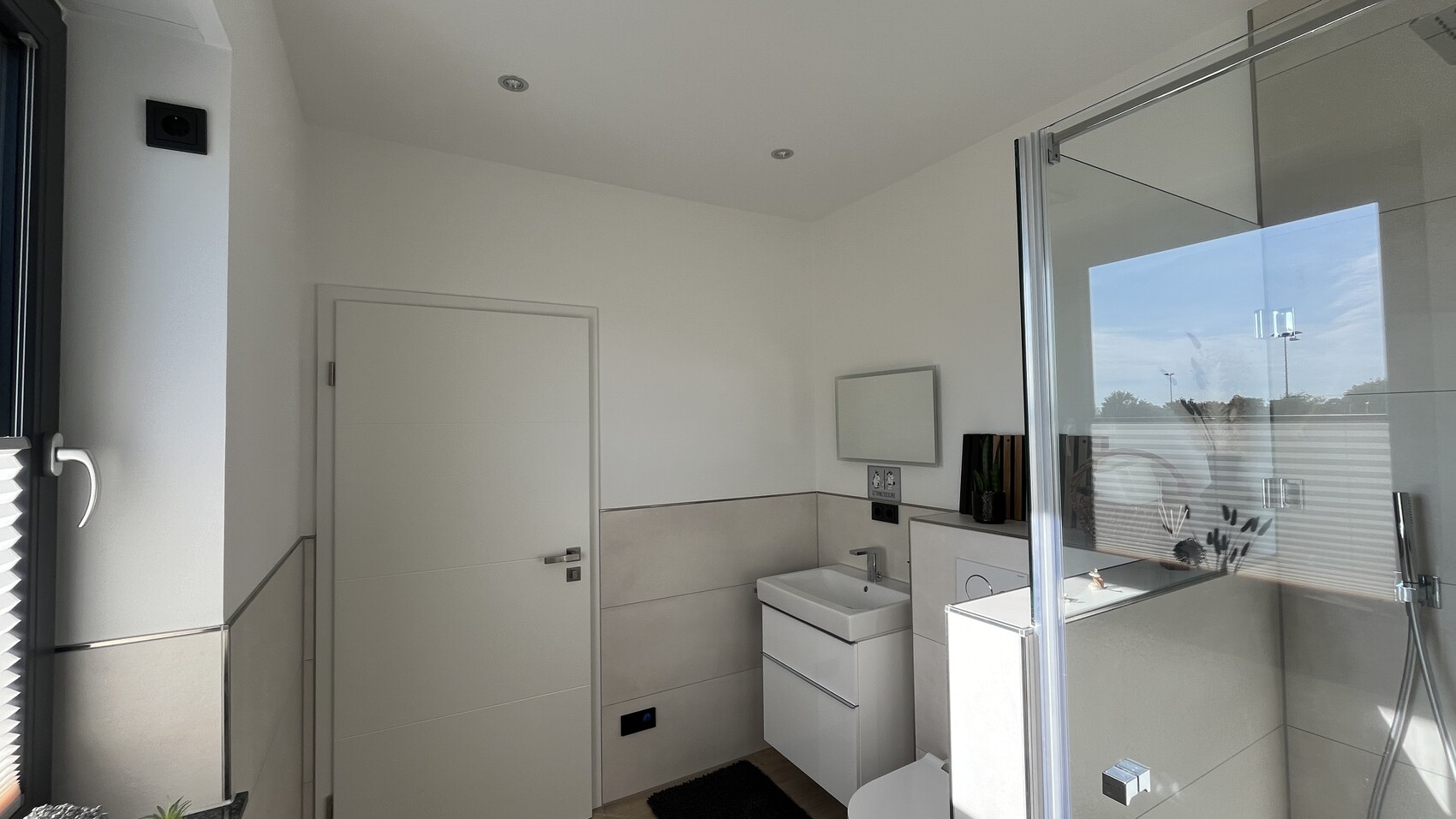 Helles Badezimmer mit Glasdusche, Waschbeckenunterschrank, Wandspiegel, Pflanze und Tür.