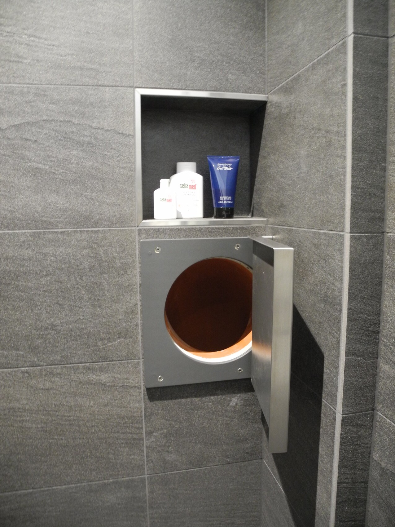 Graue Fliesenwand mit Ablage voller Pflegeprodukte; darunter eine Metallbox mit runder Öffnung.