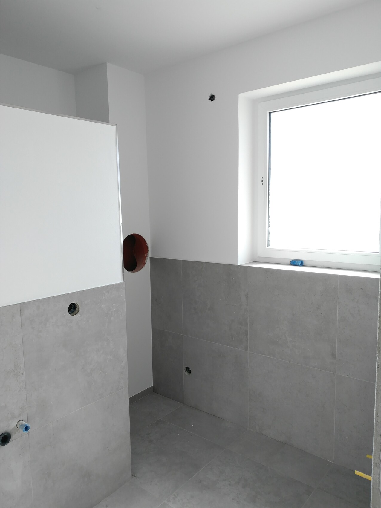 Badezimmer im Bau mit grauen Fliesen an Wand und Boden, roter Rohrdurchbruch, Fenster rechts