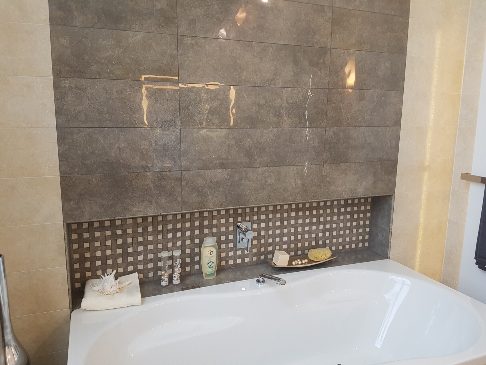 Moderne Badewanne vor grauen Fliesen und Mosaikbordüre; Seife, Lotion, Handtuch mit Muschel.