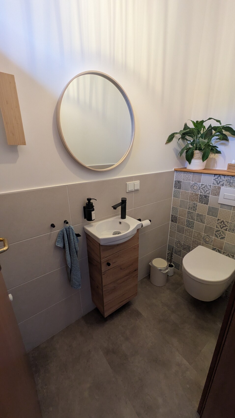 Modernes Badezimmer mit rundem Spiegel, Waschbecken auf Holzunterschrank, Toilette und Pflanze.