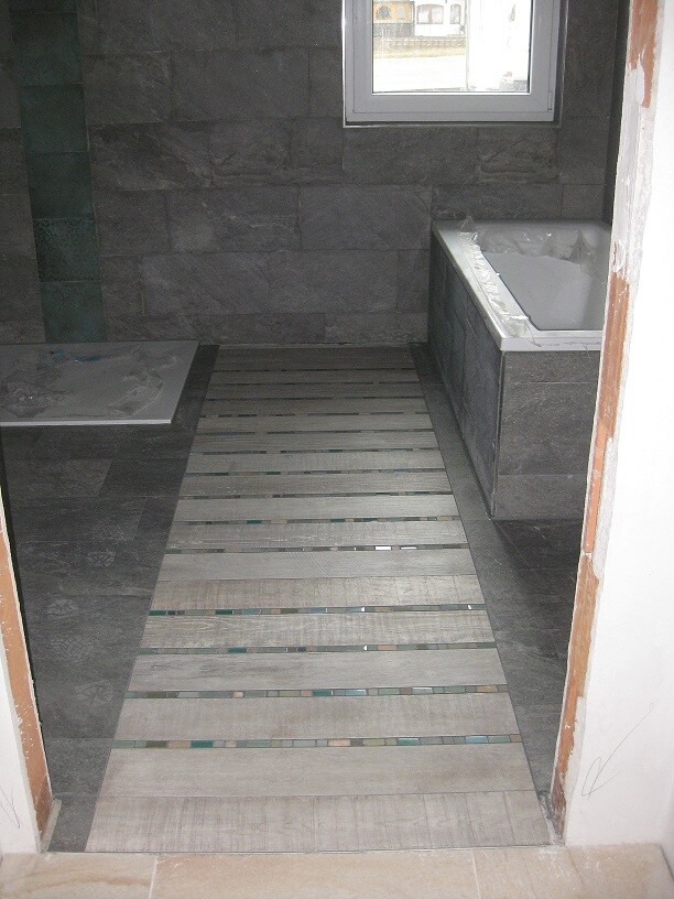 Dunkles Badezimmer mit grauen Steinwänden, Holzdielenboden und Badewanne rechts, Fenster hinten
