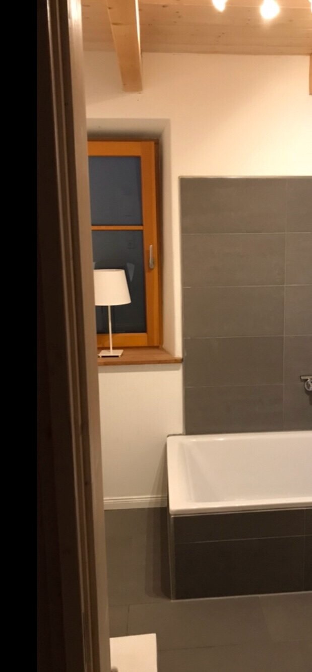 Badezimmer mit Badewanne, grauen Fliesen, Fenster mit Holzrahmen und kleinem Lampenschirm.