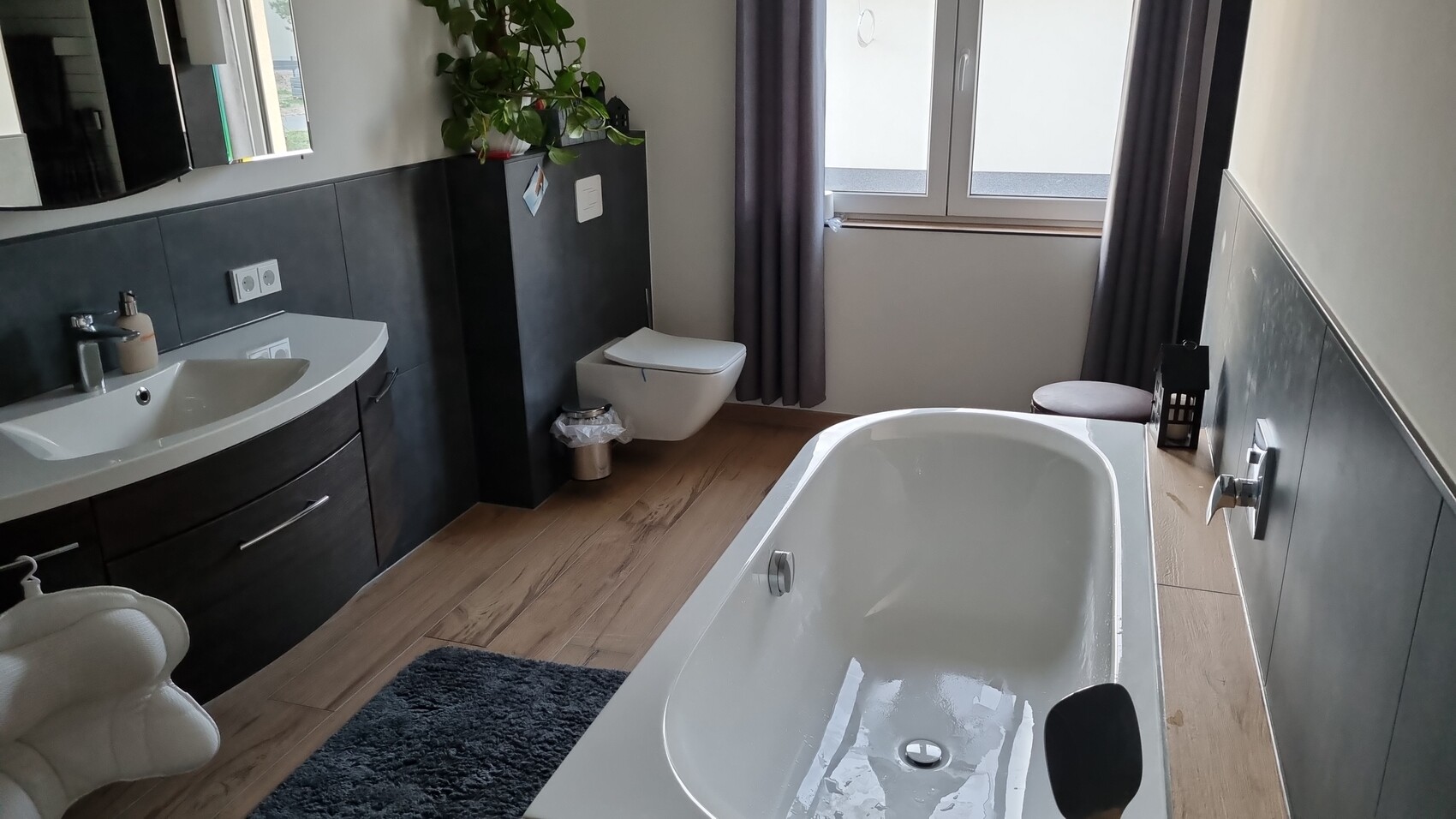 Modernes Badezimmer mit Badewanne, Waschtisch, Wandtoilette, Fenster und Pflanze.