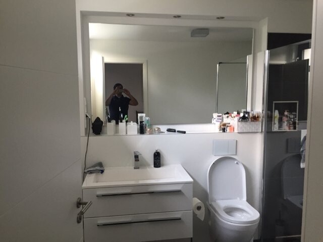 Badezimmer mit Waschtisch, Toilette und Spiegel; eine Person fotografiert sich im Spiegel.