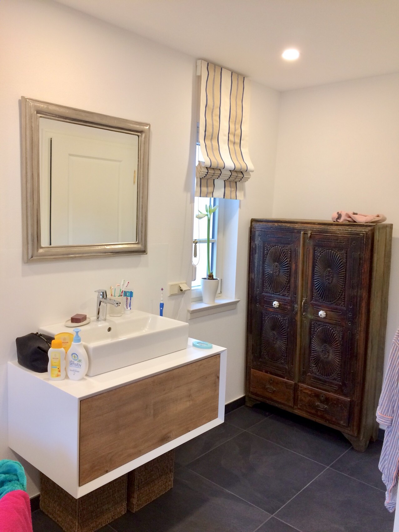 Helles Badezimmer mit Waschbecken, Spiegel, Fenster mit Streifenrollo und antikem Holzschrank.