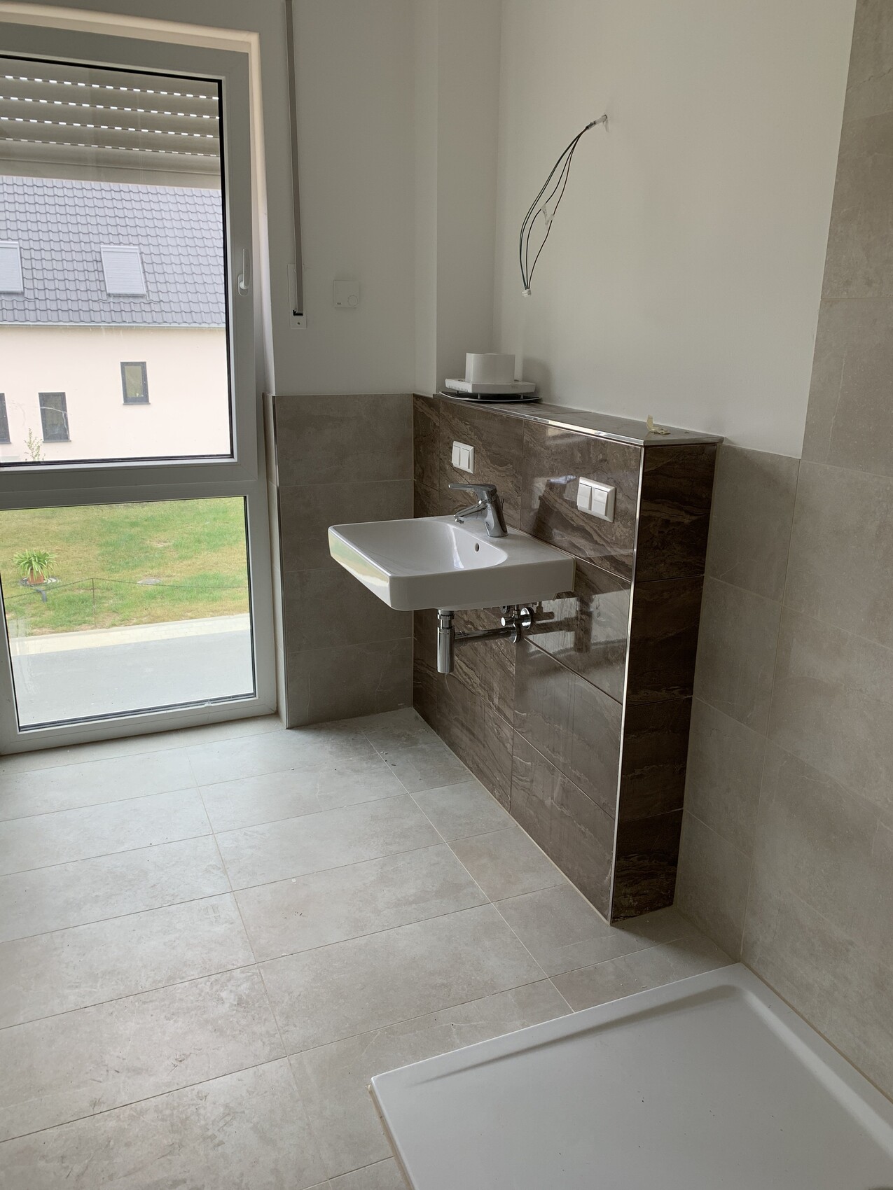 Kleines Bad mit Wandwaschbecken, braune Fliesenwand, offener Rohranschluss, Dusche im Vordergrund.